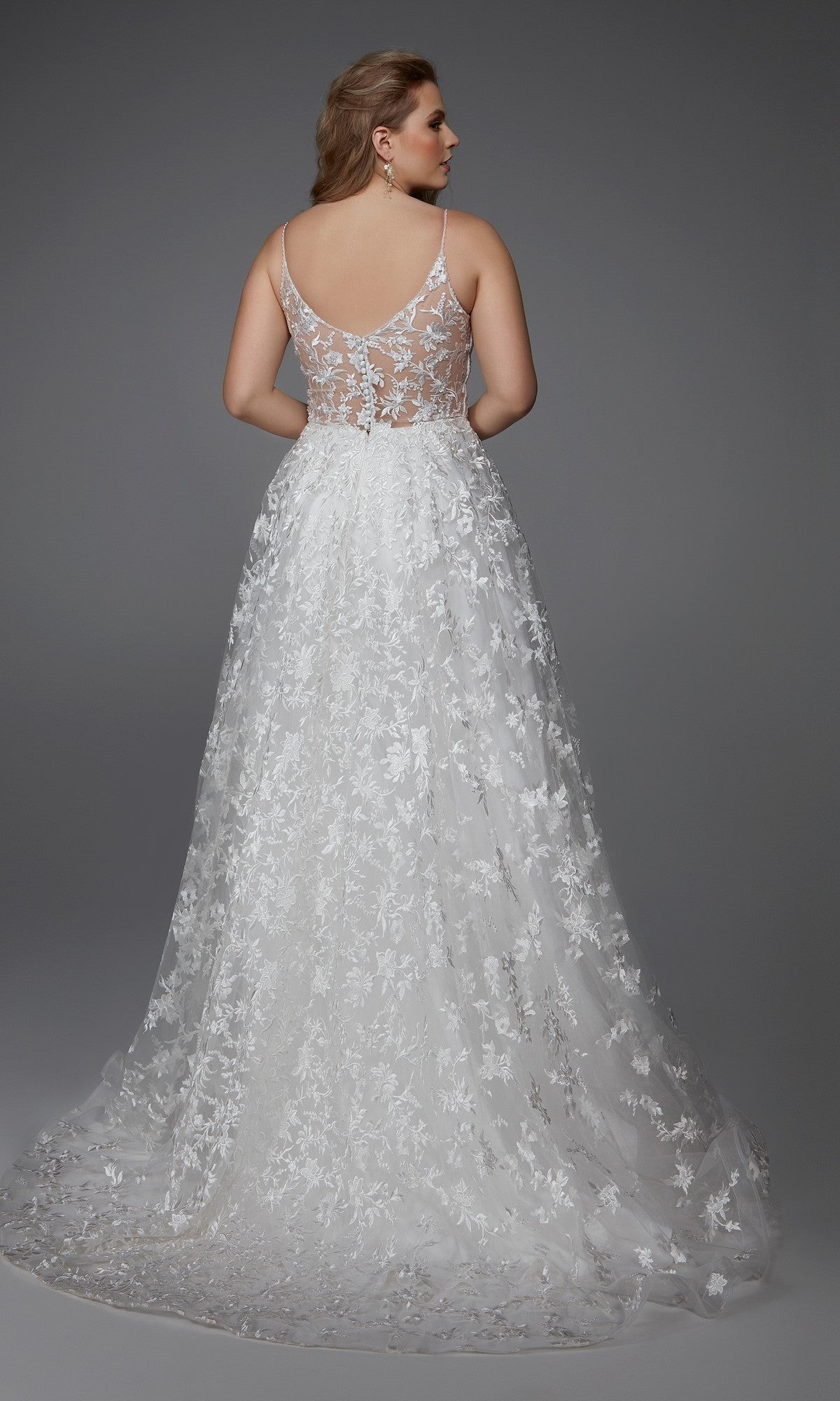 A woman faces away in the Alyce 7012 Long Formal Dress, a floor-length white gown with floral lace appliques and a sheer back, set against a plain gray backdrop.